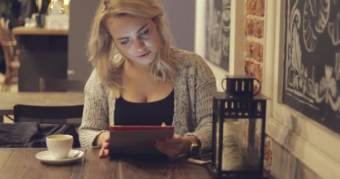 Young pretty woman sitting at table and using digital tablet while drinking cup of coffee in restaurant.