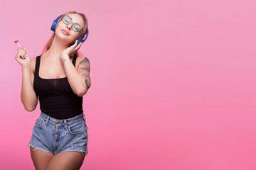 Sexy woman listening to music with her eyes closed and having a lollipop in hand. Pink background in studio photo