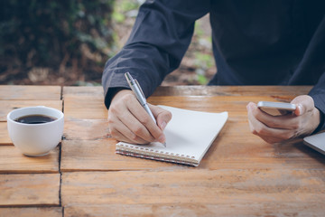 Close up businessman write notebook and using cellphone on wood table.