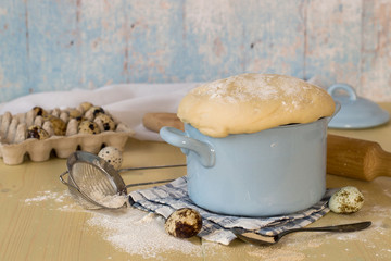 A special yeast dough for making homemade pies and buns in a small saucepan, next to quail eggs and a sieve for sifting the flour. 