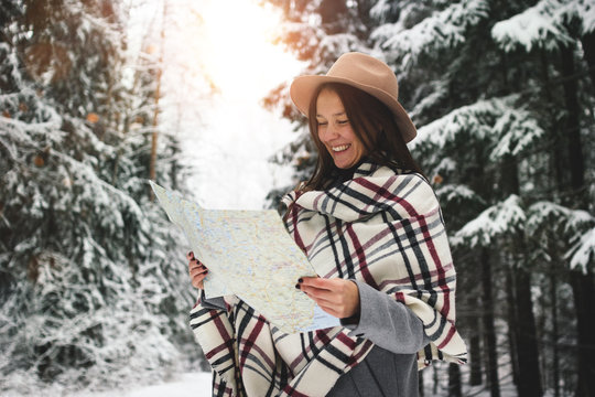 Happy and smiling traveling woman in scarf and hat hold map in hand among snowy trees in winter forest