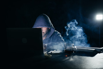 mysterious criminal man hides his face under the hood, hacks the laptop, in the dark