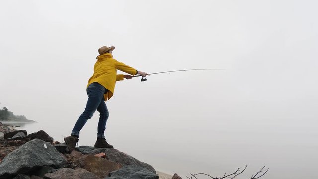 Fisherman in yellow raincoat throws fishing tackles with spinning rod in slow motion.