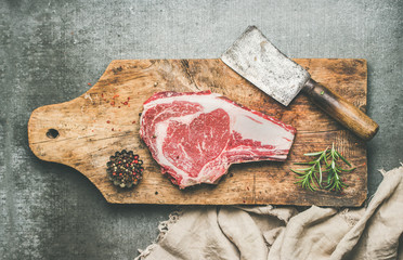 Mise à plat de viande de bœuf crue de première qualité steak de faux-filet vieilli à sec avec assaisonnement et couteau hachoir sur une planche à découper en bois sur fond de béton gris, vue de dessus. Concept de dîner riche en protéines à la viande