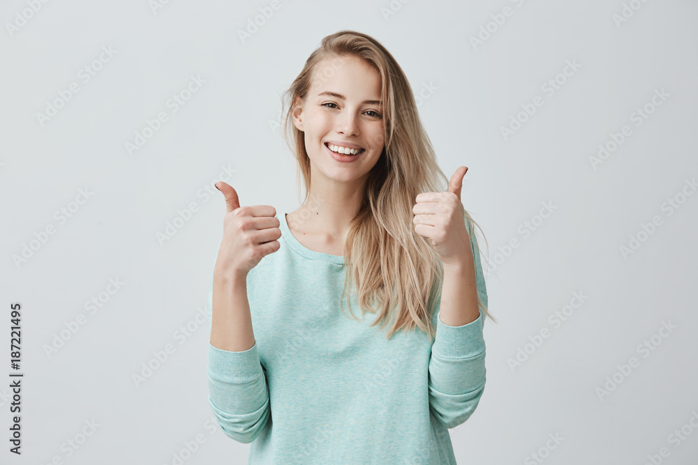 Wall mural Portrait of blonde female student or customer with broad smile, looking at the camera with happy expression, showing thumbs-up with both hands, achieving study goals. Body language - Wall murals