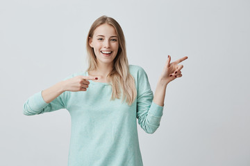 Beautiful cheerful blonde female smiling broadly at camera and pointing forefingers away, showing...