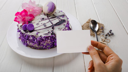 close-up, female hand holding empty blank on background cake with a flower on white wooden table . Top view