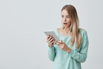 Fototapeta premium Surprised young blonde woman in long-sleeved top with wide-open mouth in shock and astonishment looking at screen of tablet, posing against gray wall background. Human face expressions and emotions