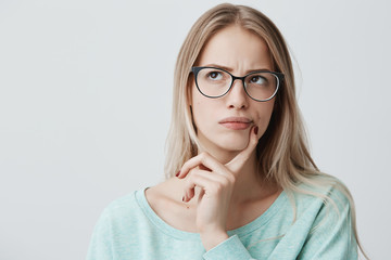 Indoor shot of thoughtful pretty woman has long blonde hair with stylish eyewear, looks aside with pensive expression, plans something on coming weekends, poses against blank wall. Puzzled female - obrazy, fototapety, plakaty