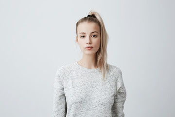Attractive beautiful female with blonde pony tail, feeling self-assuarance while posing against blank studio wall. Confident woman with dark eyes dressed casually isolated against gray background - obrazy, fototapety, plakaty