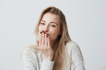 Pleasant-looking beautiful blonde female with red manicure blows kiss at camera, sends goodbye to boyfriend, poses against gray background with copy space for text or advertisment. Distant kiss