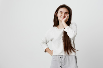 Waist-up portrait of brunette female model joyfully smiling with white teeth, glad to receive good news or present. Excited girl expresses positive emotinos, holds hand on cheek in amazement