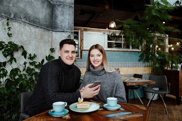 Young beautiful loving couple in cafe. The man and the woman watch a photo on the smartphone, embrace, have tasty tea and eat a dessert. 