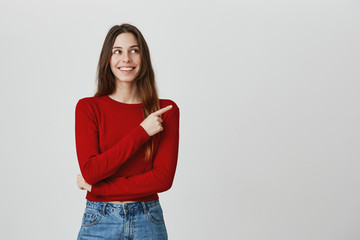 Copy space for advertisement. Positive emotions. Studio portrait of young attractive caucasian girl with long hairstyle in stylish hip outfit smiling, pointing aside with happy face expression.