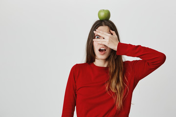 Copy space. Healthy lifestyle. Close up portrait of young beautiful caucasian girl with long hair in red jumper looking in camera, closing eyes with scared expression, holding apple on head.