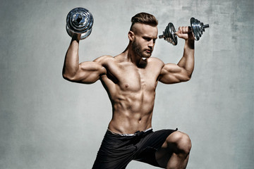 Young male bodybuilder doing exercise with heavy weight dumbbells. Photo of muscular man on grey...