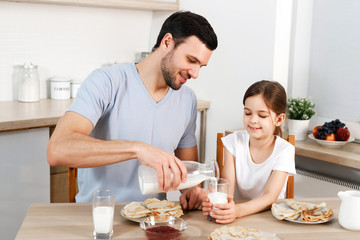 Obraz na płótnie Canvas Happy father and daughter have breakfast at kitchen, eats delicious pancakes with jam, drink milk, enjoys delicious food prepared by mother. Family apetizes yummy sweet dish. Eating and people concept