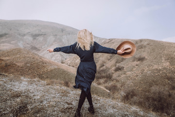 stylish blond girl in blue dress and with beige hat in hands is standing with her back in the mountains on nature with beautiful view and scandinavian landscape - 187240215