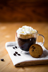 Morning coffee with cookie, whipped cream topping on beige napkin in double botton glass on wooden brown table background