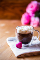 Valentine`s day celebration concept. Cup of coffee with cream froth on brown napkin in double bottom glasses with heart love shape sugar on wooden background with pink flowers