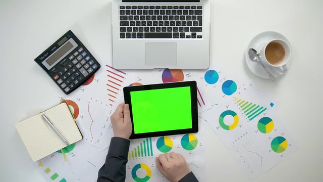 A man's hands holding an i-Pad and touching the green sreen over a white table with diagram sheets. View from the top. Close-up shot.