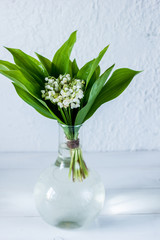Bouquet of lily of the valley in a little transparent vase on wooden light background. Fresh spring mood