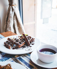 a beautifully decorated wooden table by the window with pastries and a waffle with berry sauce or chocolate with a cup of tea