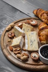 Assorted cheeses on round wooden board plate Camembert or Brie cheese, walnuts, croissant, bread, fruits and berries, dark black wood background, top view.