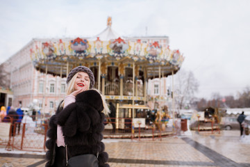 Adorable smiling model wears knitted hat and fur coat, posing near the carousel