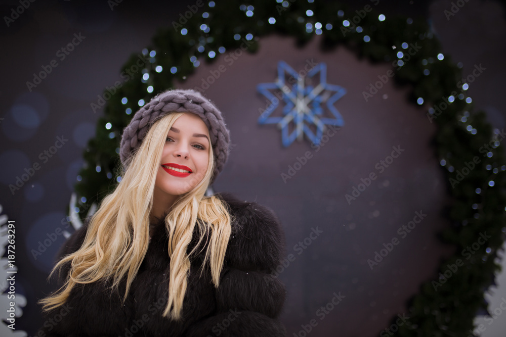 Wall mural street portrait of emotional young woman wearing knitted hat and fur coat, posing at the christmas f