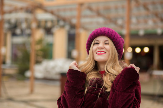 Funny young model wearing red knitted hat and winter coat, posing on the background of garland at the street in Kiev