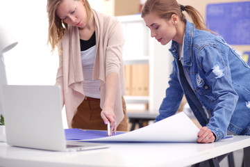 Obraz premium Two young woman standing near desk with instruments, plan and laptop.