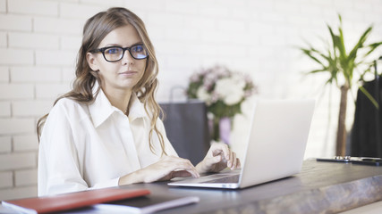 A young beautiful woman wearing glasses typing at her laptop in office and looking at camera.