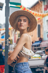 A beautiful young blonde woman exploring pretty part of Chinatown in Singapore. Hot weather in the colorful small urban downtown. She wears casual combination of clothes with a nice hat.