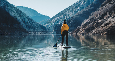 a woman with yellow plumber paddle surfing on a lake in a snowy mountain landscape - obrazy, fototapety, plakaty