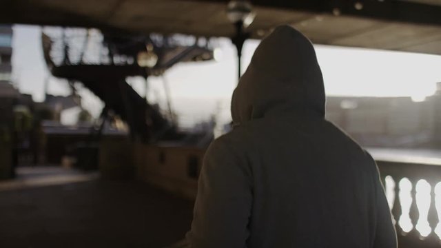 Camera follows a hooded jogger as they run by a river in the city, in slow motion