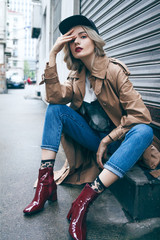A young beautiful blonde woman is sitting and enjoying small urban street in Shanghai. She is wearing nice long brown coat and black hat. Her gorgeous face is accentuated by a dark red lipstick.
