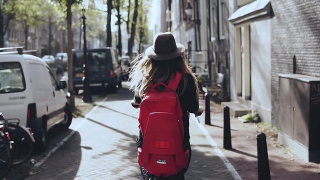 Back view. Female on a sunny city bicycle ride. Slow motion. Girl in stylish hat with red backpack on bike. Exploring.