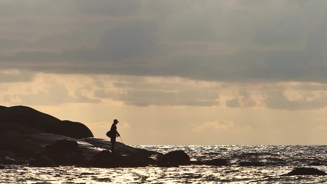 Slow motion shot of  a silhouette of fisherman fishing on rocks against golden sunset water with waves and splashes with beautiful cloudy background