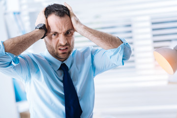 Awful. Young bearded man standing in his light office and pressing his head with two hands and showing his anger while looking in front of himself