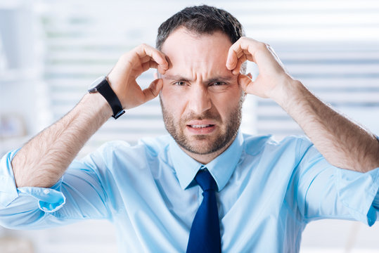 How terrible. Expressive irritated young employee feeling upset and angry while standing in his office after a difficult day and touching his head with both hands