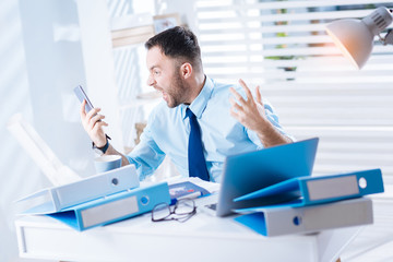 Irritated. Emotional young irritated worker sitting at the table covered with folders and looking...
