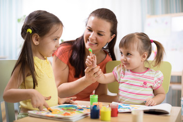 Kids with young teacher woman painting on table together in kindergarten .