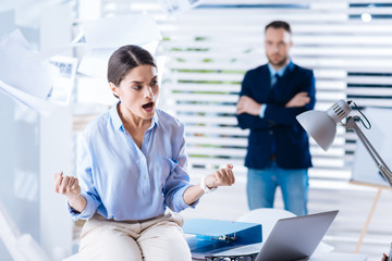 Furious woman. Emotional angry young woman sitting on the table and clenching her fists while shouting while her calm colleague standing behind her back with his arms crossed