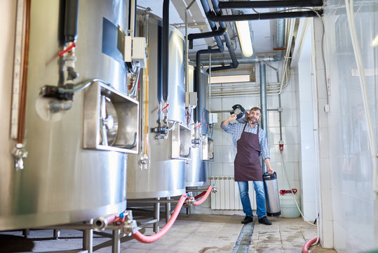 Full length portrait of cheerful middle-aged worker wearing apron looking away with toothy smile while carrying heavy beer kegs and walking along corridor of modern brewery.
