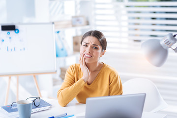 Terrible toothache. Young exhausted worker sitting at the table and touching her cheek while suffering from the unbearable toothache