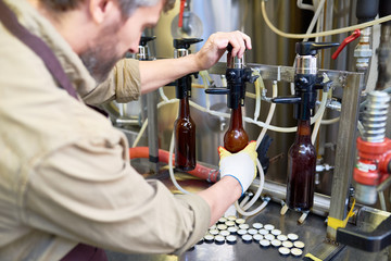 Profile view of bearded machine operator wearing gloves and apron filling beer bottles from tank...