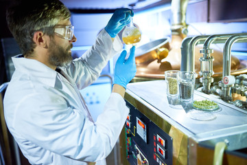 Confident inspector wearing white coat and protective goggles holding flask with beer in hands while checking its purity