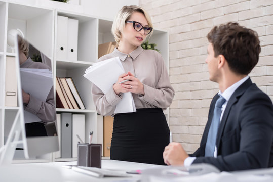 Portrait Of Pretty Blonde Businesswoman Talking To Boss Standing By Desk In Modern Office And Holding Heap Of Papers, Copy Space