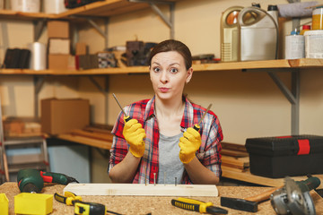 Beautiful caucasian young woman in plaid shirt, gray T-shirt, yellow gloves twisting by screwdriver screw, working in carpentry workshop at wooden table place with piece of wood, different tools.